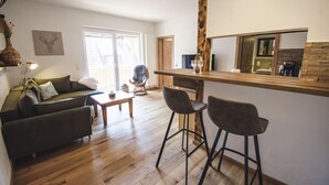 Ein modernes Wohnzimmer mit Holzboden, einer Couch, zwei Barhocker, einem Couchtisch und einem Esstisch mit Flasche und Gläsern.