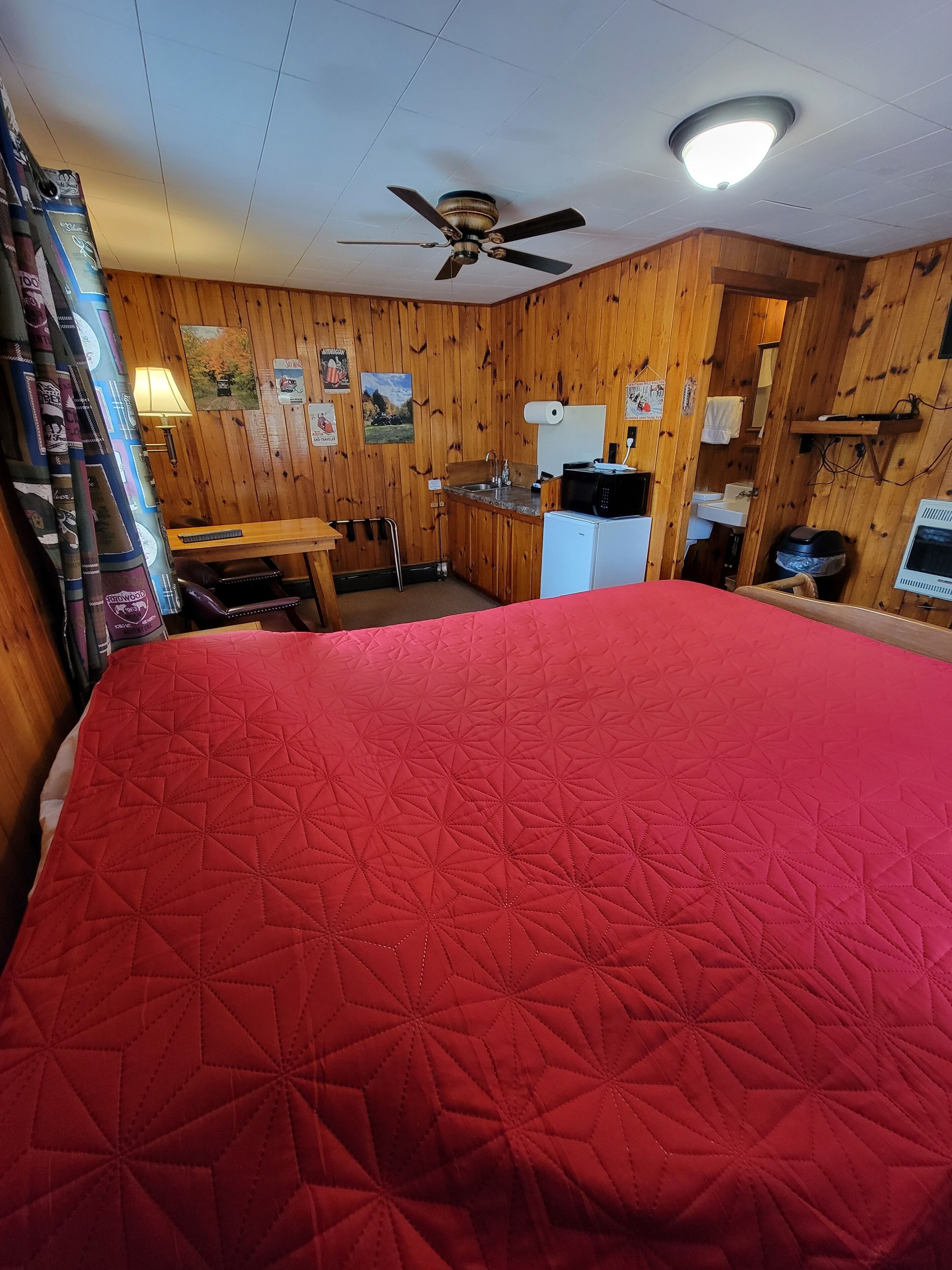 A room with a large bed, wooden walls, a ceiling fan, a small kitchenette, and a window with curtains.