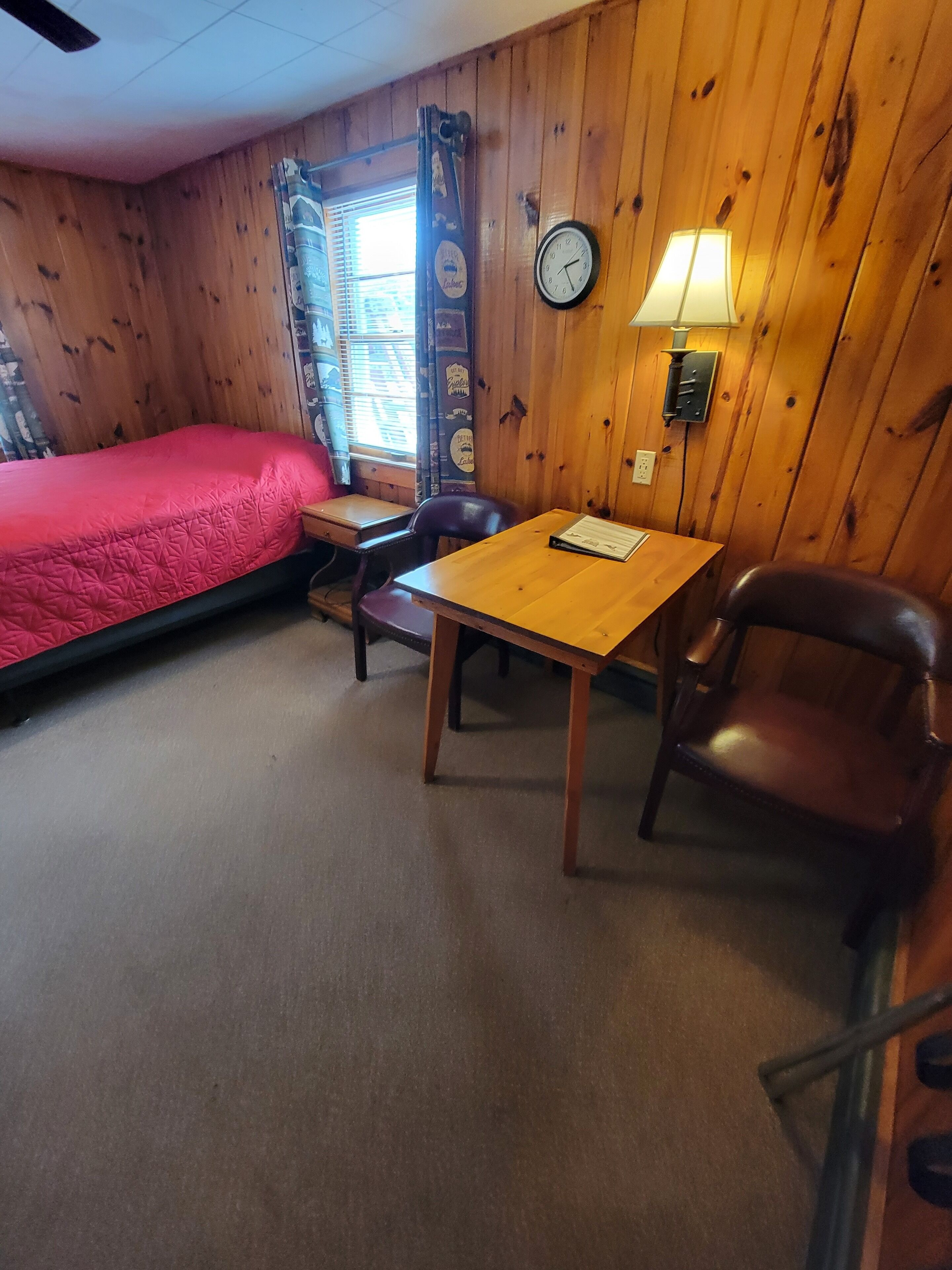 Une pièce aux murs en bois, un lit recouvert d’une couverture rouge, une table avec une lampe, une chaise et une horloge au mur.