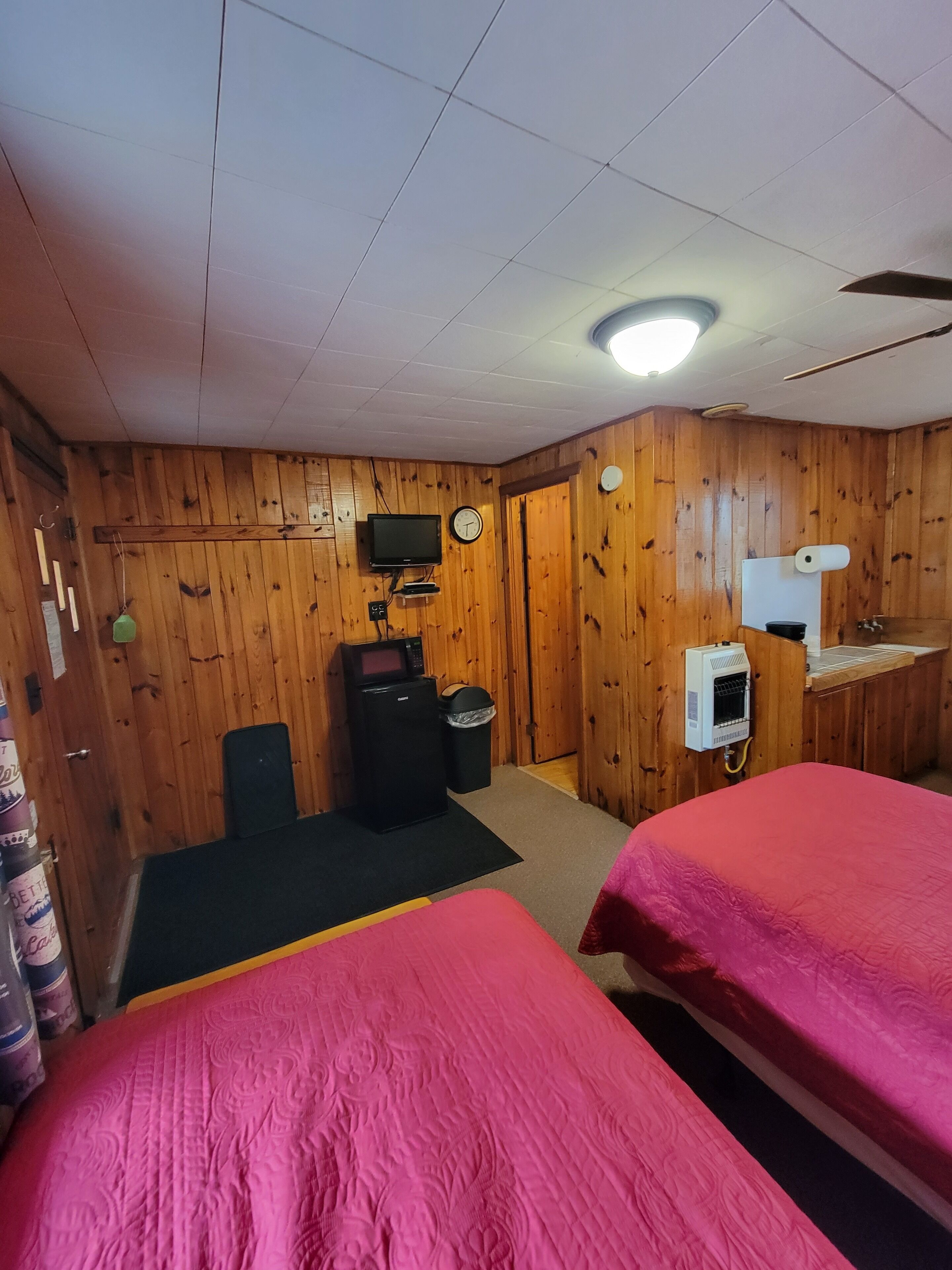 A room with wooden walls, two beds with pink bedding, a television, a microwave, a heater, and a trash bin.
