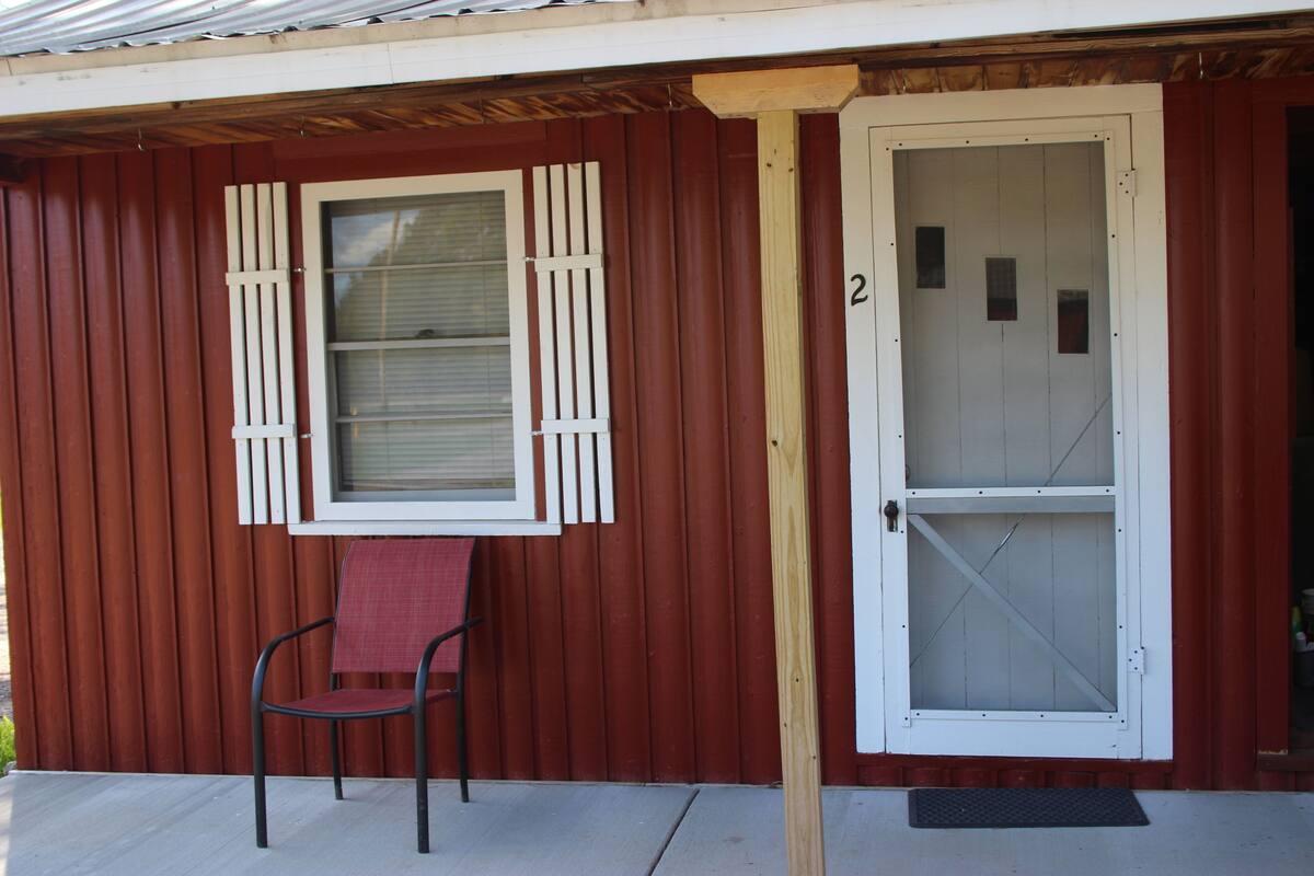 A wooden building with a red exterior, a white door with a glass panel, and a single chair with a red cushion outside.