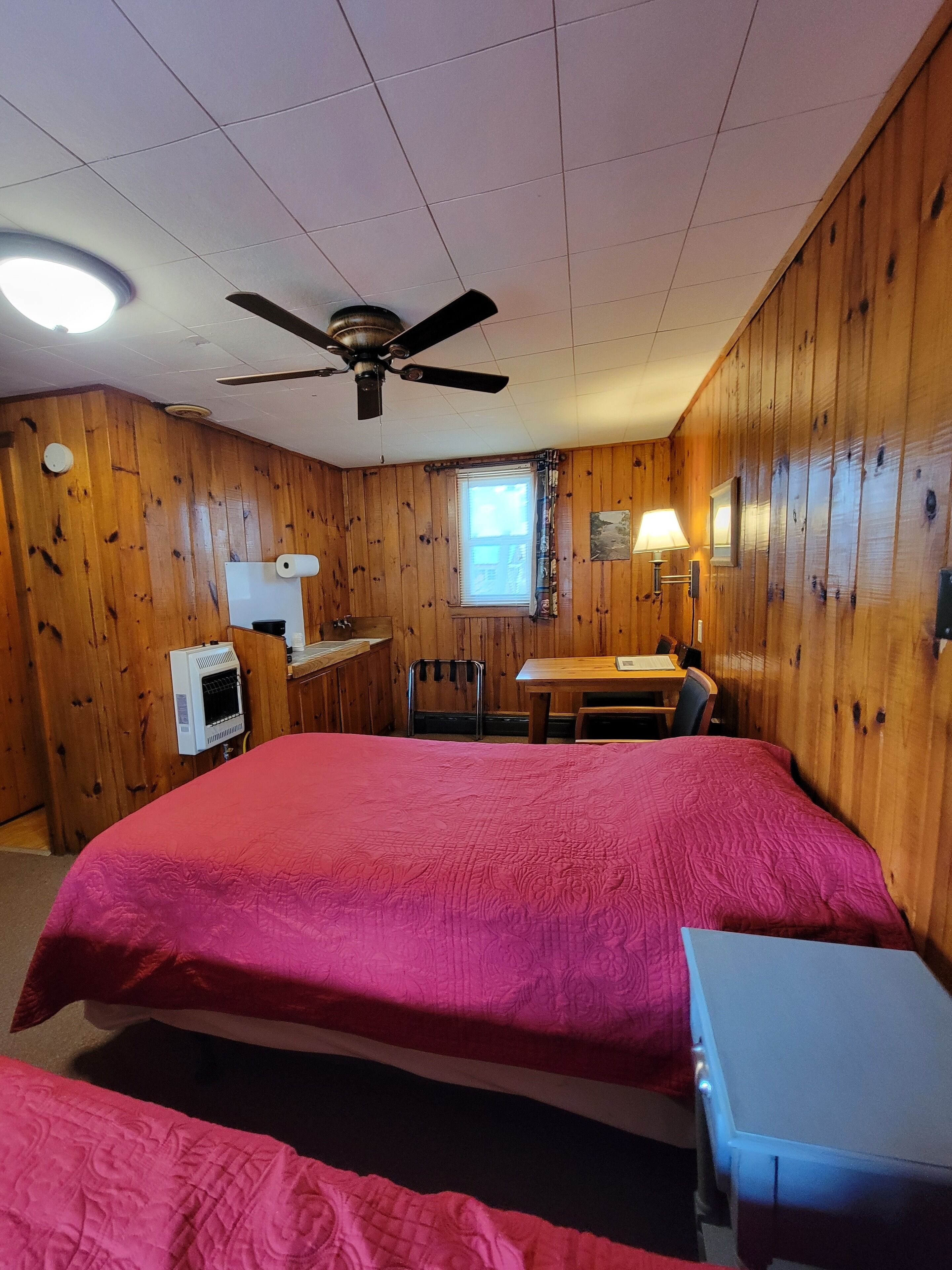 Een kamer met houten wanden, een plafondventilator, een bed met een rode sprei, een klein tafeltje en een raam met jaloezieën.