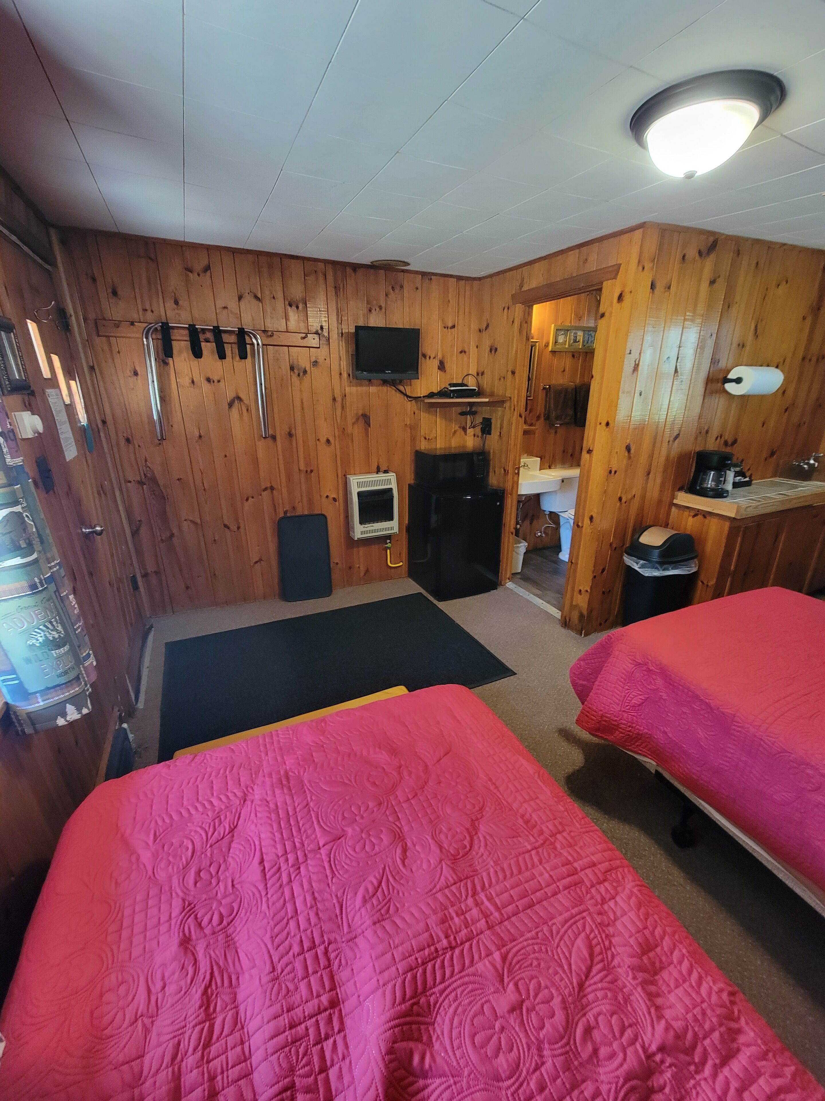 A room with wooden walls, two beds with red bedding, a television, a microwave, and a small kitchenette.