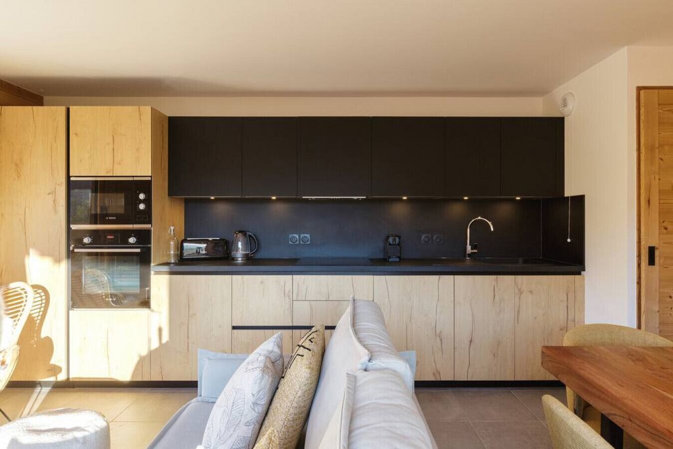 A modern kitchen with dark cabinets, a built-in oven, a toaster, and a coffee maker. There is a wooden dining table and a sofa with cushions.