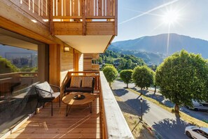A wooden balcony with a sofa and table, overlooking a road and trees with a mountain in the background.