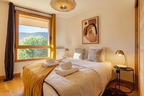 Een slaapkamer met een groot raam, een bed met witte lakens, een schilderij aan de muur en een nachtkastje met een lamp.