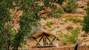 A rustic cabin with a stone foundation, surrounded by greenery and a body of water. - Cave Lakes Canvas Cabin Suite with Soaker tub #2 (Kanab)