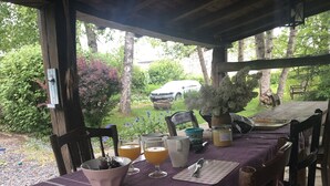 A covered outdoor dining area with a table set for two, featuring a purple tablecloth, a bowl of fruit, and a vase of flowers. - Charming country house with large barbecue (Gouvy)