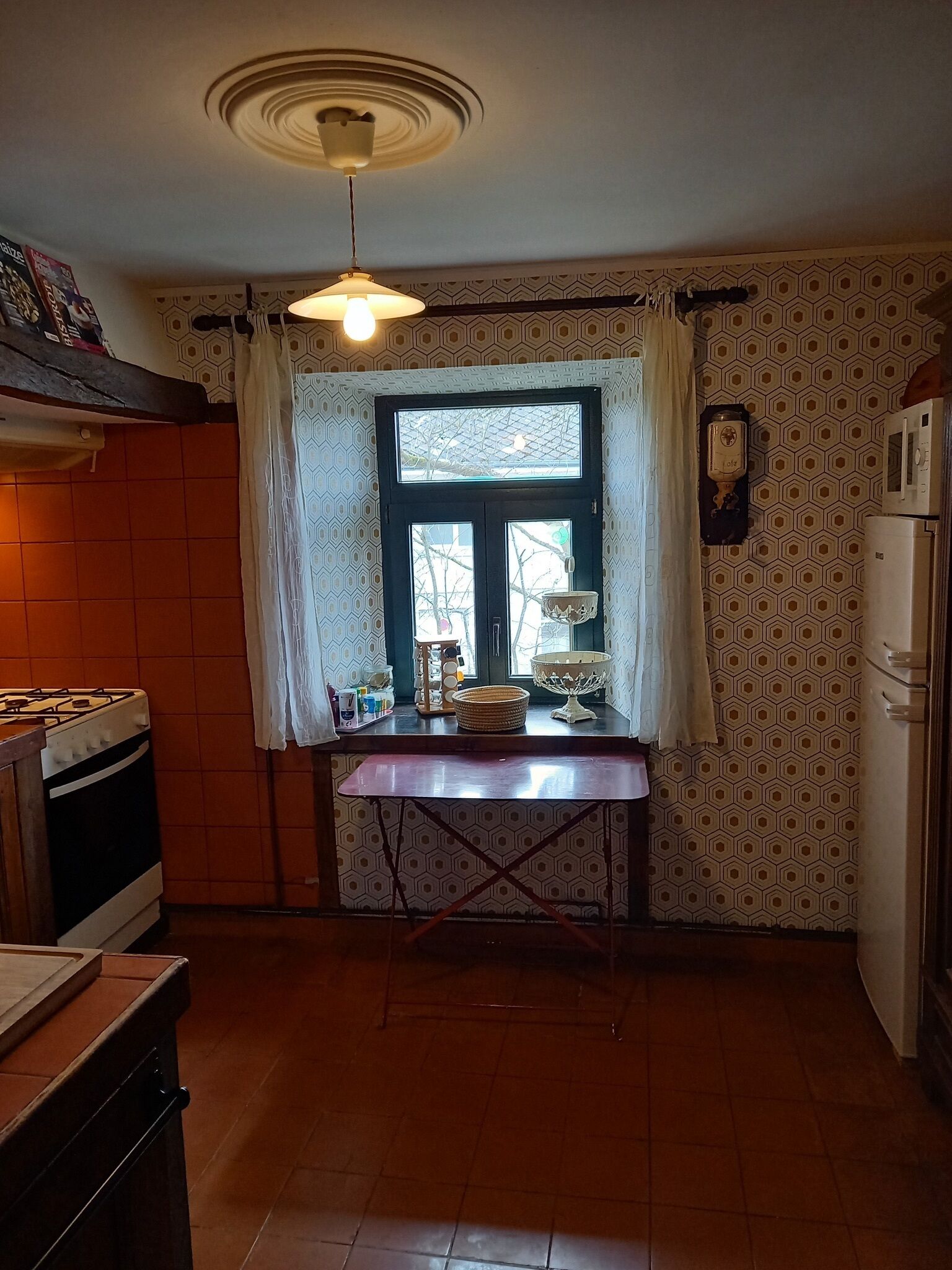 A kitchen with a gas stove, a refrigerator, a window with curtains, and a hanging light fixture.