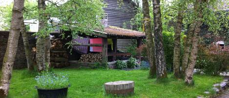Ein rustikales Holzhaus mit Strohdach, umgeben von einem üppigen Garten mit einer Steinmauer, einer Holzbank und einem großen Pflanzgefäß mit blauen Blumen.