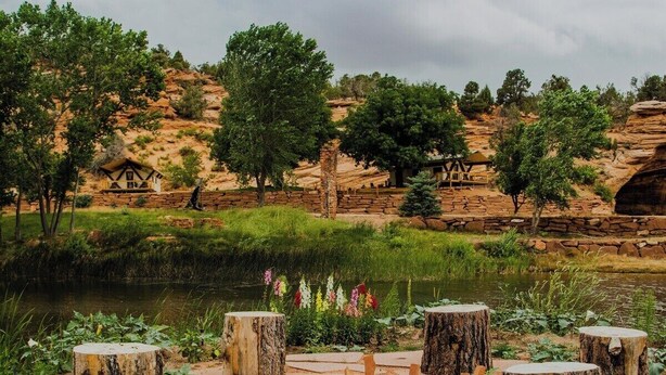 Un espace extérieur rustique aménagé avec des bancs en pierre et un foyer, entouré d’un paysage naturel composé d’arbres et d’une étendue d’eau.