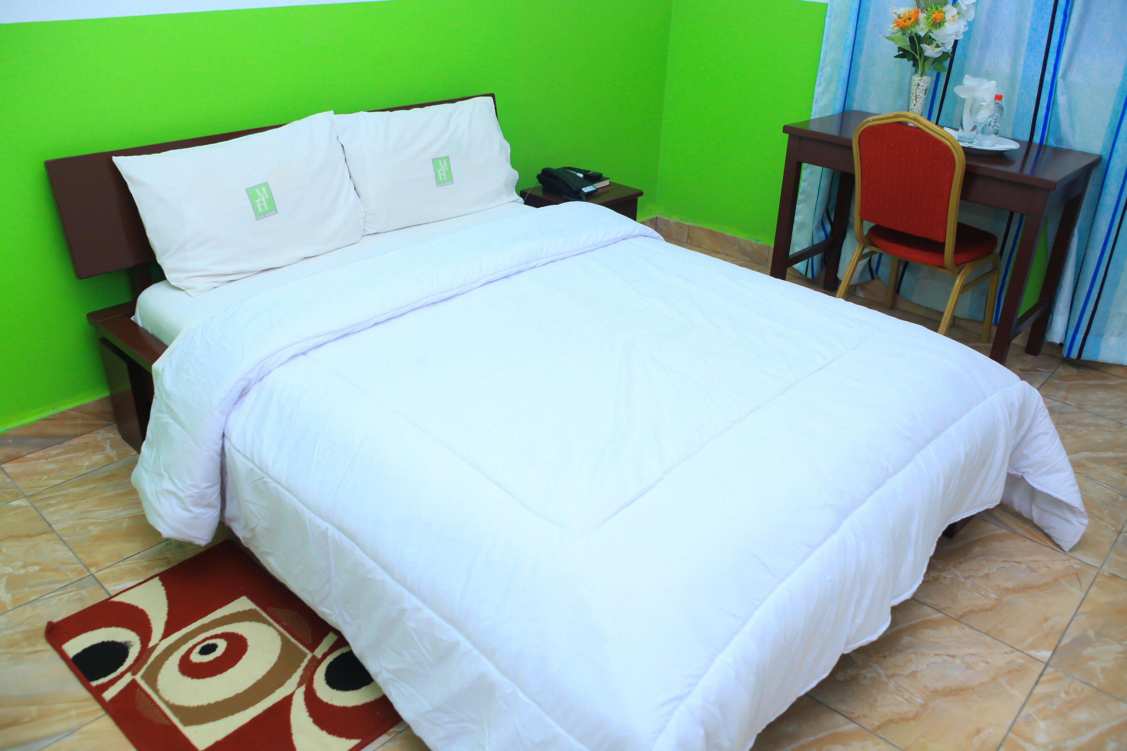 Una cama bien hecha con ropa de cama blanca, una mesita de noche con un teléfono y un libro, un escritorio con una silla, y un florero con flores.