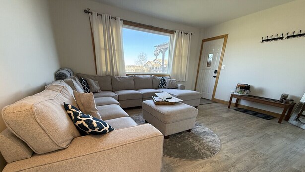Ein Wohnzimmer mit beiger Couch, Hocker und Couchtisch. Ein Fenster mit Vorhängen, eine Tür und ein Garderobenschrank sind an der Wand angebracht.