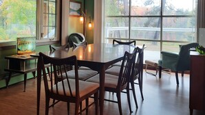 A dining area with a wooden table and chairs, large windows with a view of trees, and an air conditioning unit on the wall. - Luxury Winter Retreat/Direct Snowmobile Access (Spurr Township)