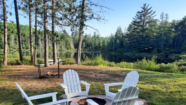 A circular fire pit with four white Adirondack chairs around it, set on a brick patio with a forested background. - Luxury Winter Retreat/Direct Snowmobile Access (Spurr Township)