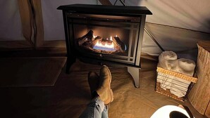 Eine Person sitzt auf dem Boden vor einem Holzofen, mit einem Wäschekorb voller Handtücher und einer Stehlampe.