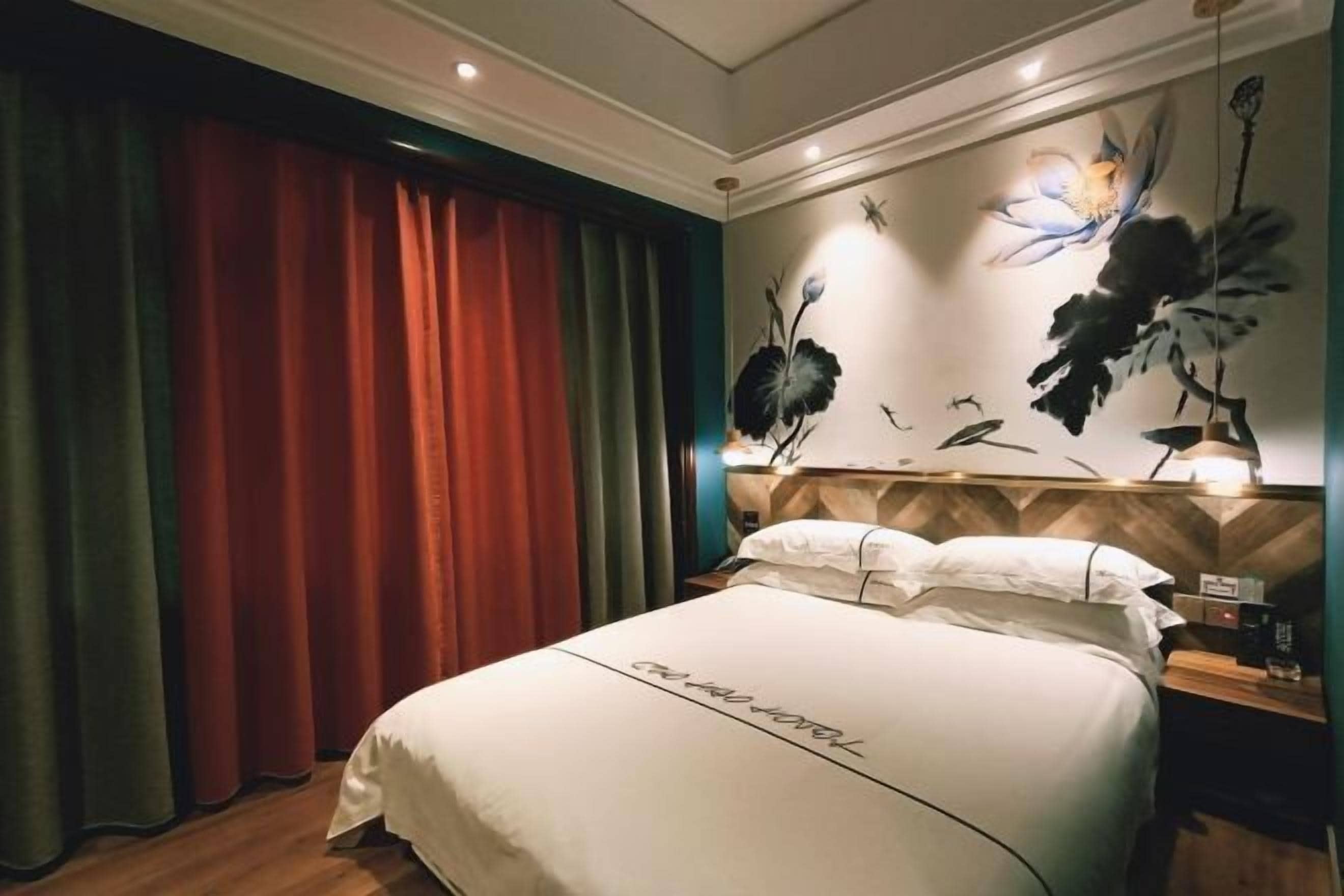 A hotel room with a bed, curtains, a wall with floral artwork, and a nightstand with a lamp.