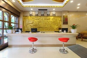 Un área de recepción con mostrador, dos taburetes rojos, un muro decorativo con relieves dorados y un ventanal grande a la izquierda.