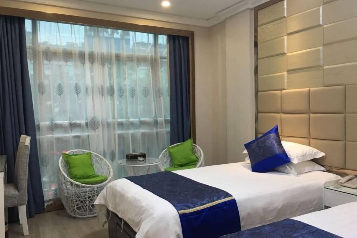 Une chambre d’hôtel avec deux lits, une petite table, deux chaises et une grande fenêtre avec des rideaux.