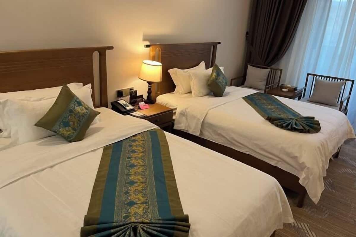Habitación de hotel con dos camas, una mesita de noche con lámpara, una silla y una ventana con cortinas.