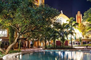 Zona de piscina con un edificio grande al fondo, rodeada de árboles y plantas de palma.