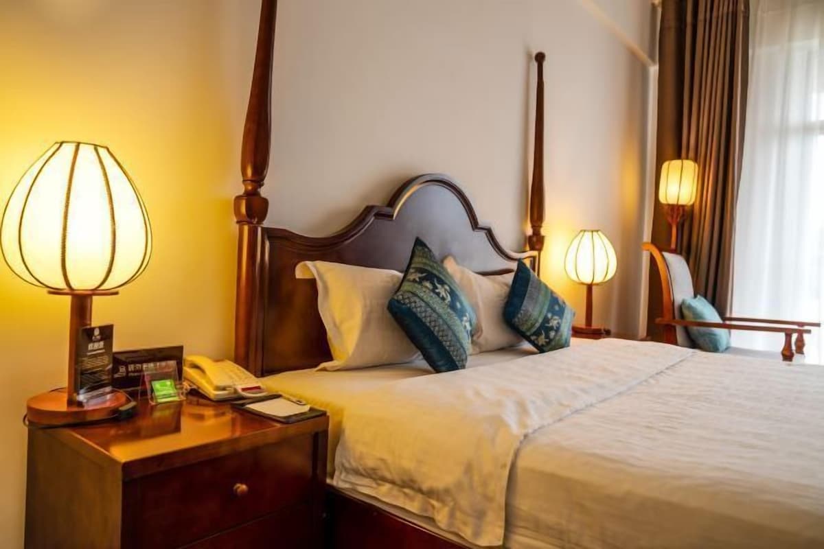 A hotel room with a four-poster bed, bedside lamps, a nightstand with a telephone and a card, and a chair with a lamp.