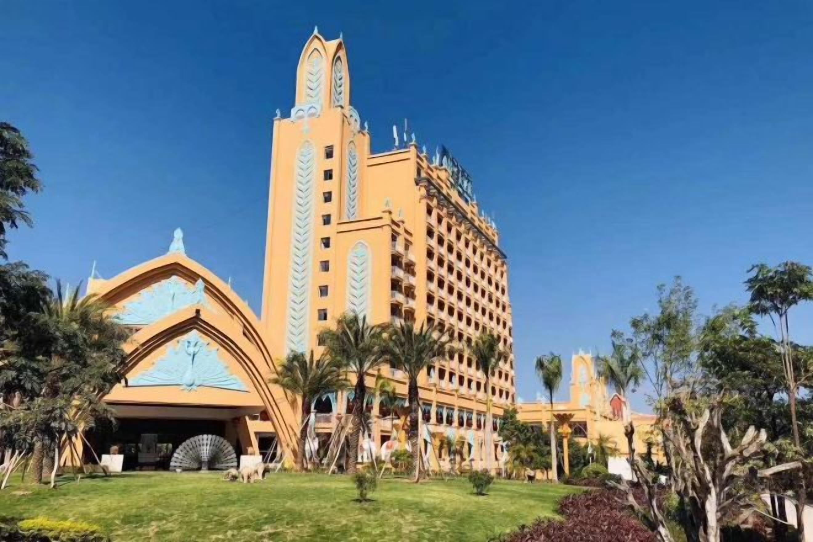 A hotel with a prominent clock tower, a large central building, and a well-maintained lawn with palm trees.