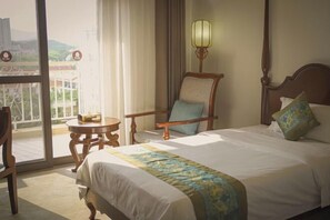A hotel room with a bed, a chair, a small table, a lamp, and a view through a balcony door.