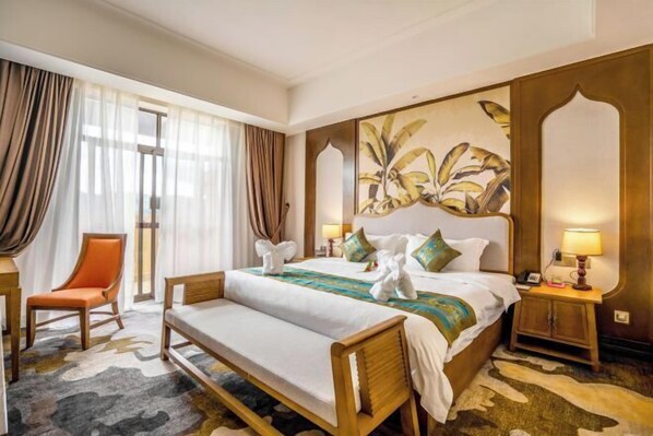 A hotel room with a large bed, a wooden bench, a chair, and a nightstand with a lamp.