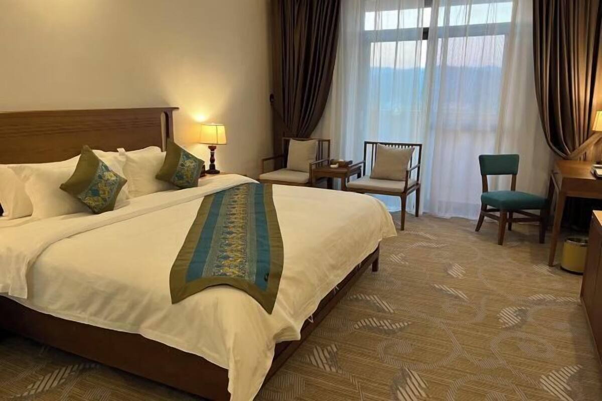Habitación de hotel con una cama grande, dos sillas, un escritorio y vistas al exterior a través de cortinas translúcidas.