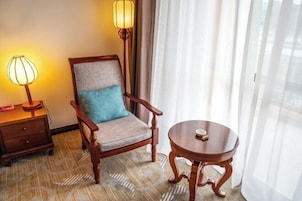 Quarto de hotel com uma cadeira de madeira, uma pequena mesa redonda, uma luminária e uma janela com cortinas leves.