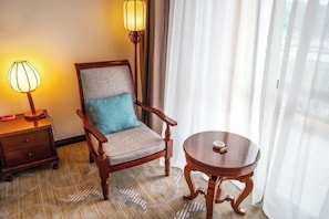 A hotel room with a wooden chair, a small round table, a lamp, and a window with sheer curtains.