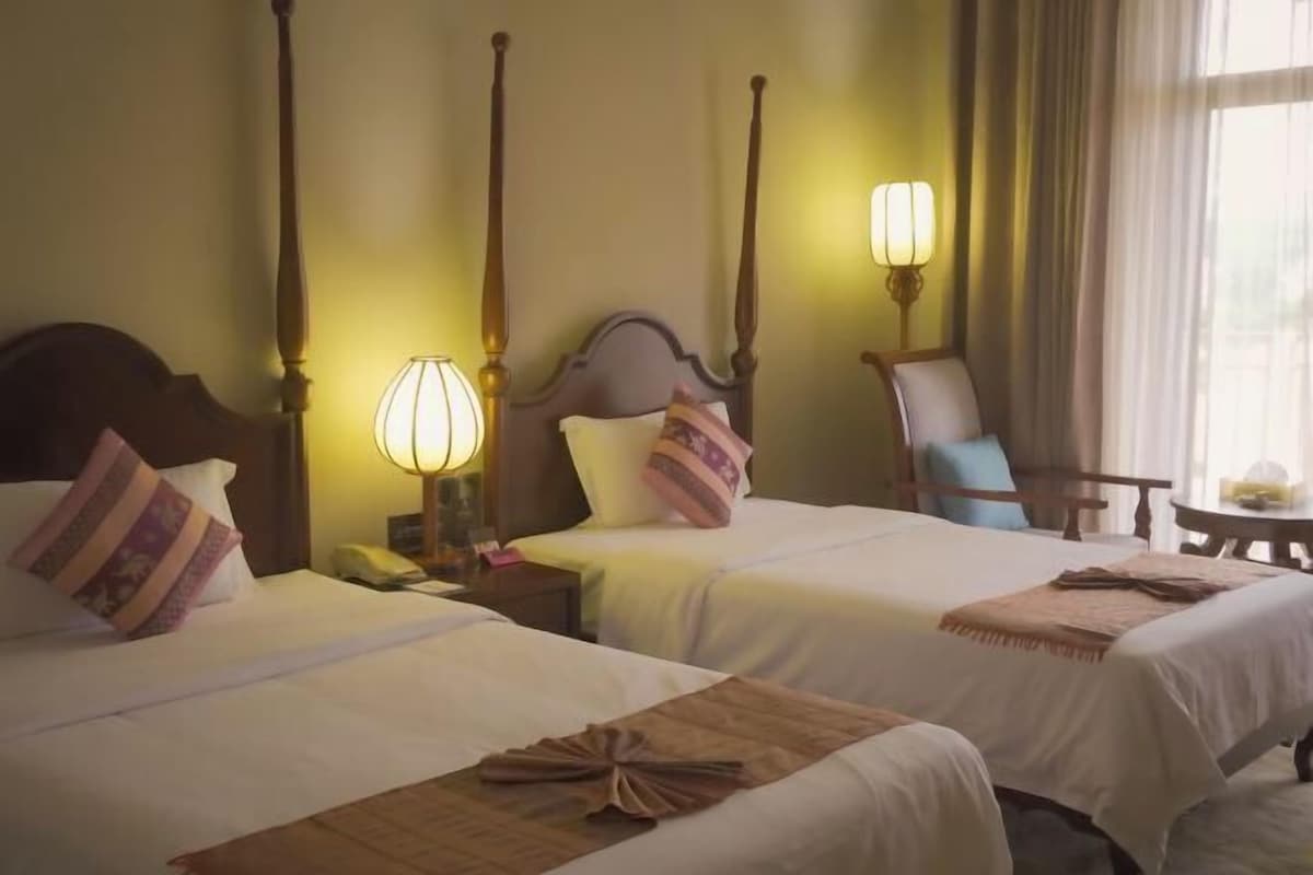 A hotel room with two beds, a nightstand, a lamp, a chair, and a small table.