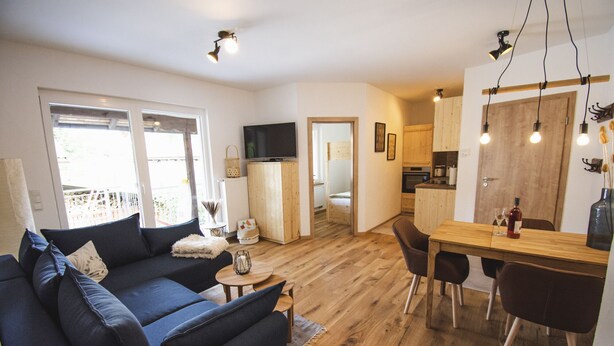 A modern living room with a dark blue sofa, a wooden dining table with chairs, and a kitchen area in the background.