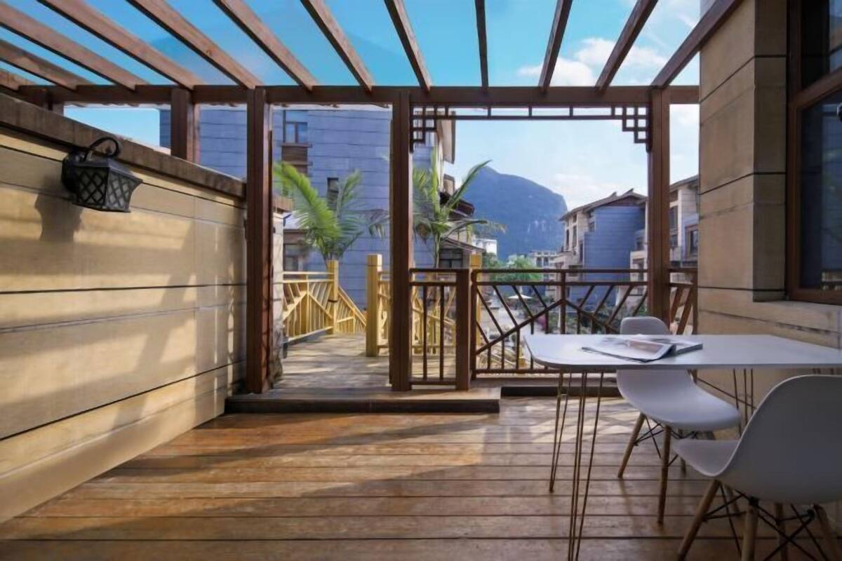 Ein überdachter Außenbereich mit Holzboden, einem Tisch mit Stühlen, einer Laterne und Blick auf Gebäude und Berge.