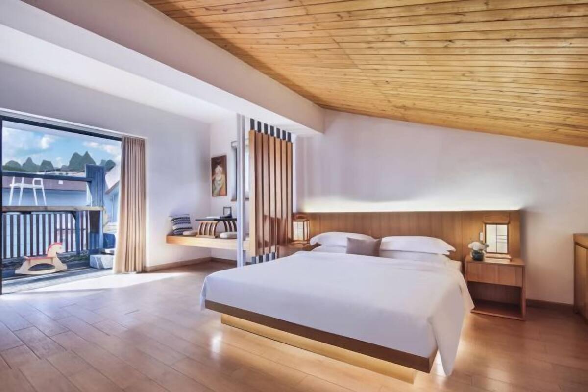 Ein modernes Schlafzimmer mit einem großen Bett, einer Holzbalkendecke, Blick ins Freie, einem Nachttisch und einem Beistelltisch.