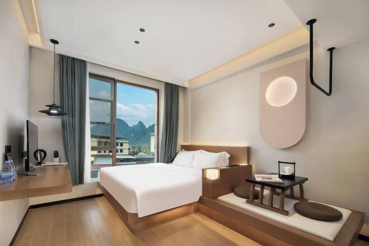 Ein modernes Hotelzimmer mit einem großen Bett, einem Schreibtisch mit Computer, einem Nachttisch mit Lampe und Blick auf Berge durch das Fenster.