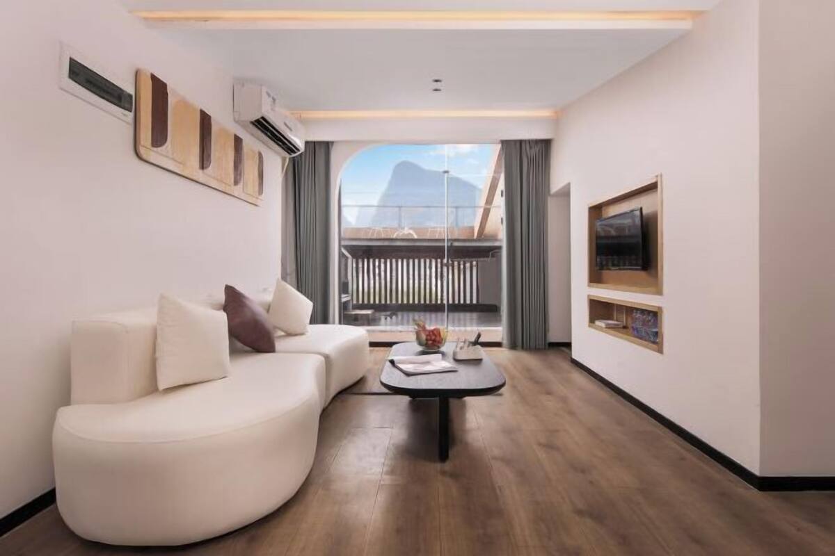 Ein modernes Hotelzimmer mit einer gebogenen Couch, einem kleinen Couchtisch, einem Fernseher und einem Balkon mit Bergblick.