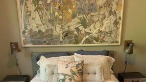 A bedroom with a large bed, two nightstands, a wall-mounted lamp, and a large painting above the bed.
