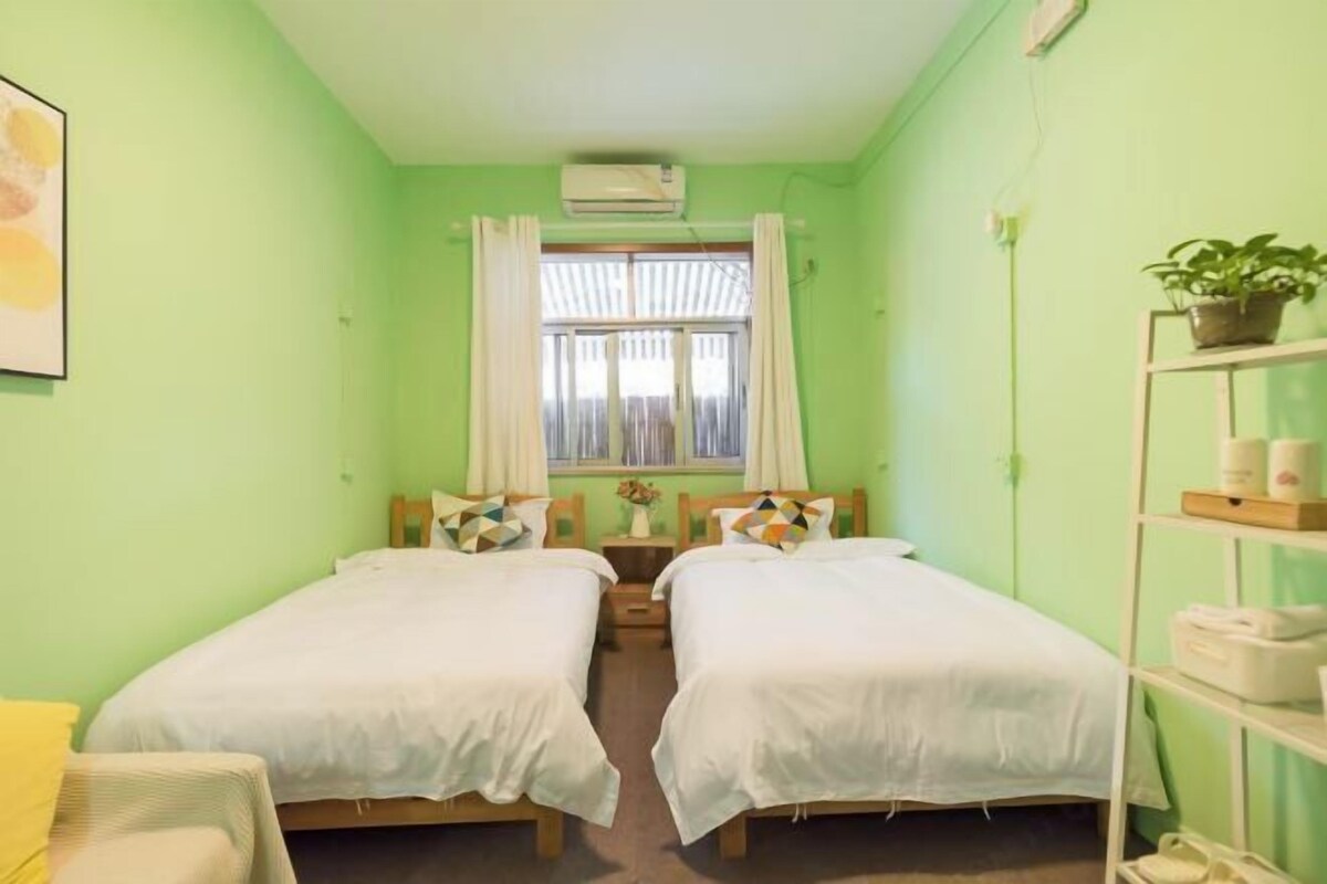 Quarto com duas camas, janela com cortinas, uma pequena mesa com um vaso e uma prateleira com objetos.