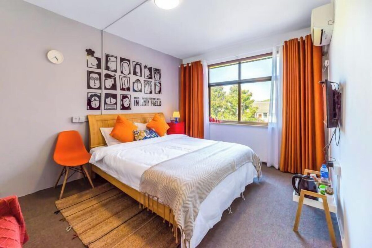 Quarto com cama, cadeira laranja, relógio na parede e janela com cortinas.