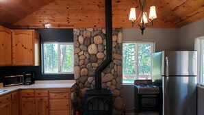 Una cocina con armarios de madera, pared de piedra, estufa a leña y refrigerador de acero inoxidable, además de una ventana con vista a árboles.