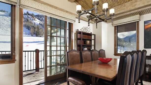A dining room with a large wooden table, leather chairs, and a chandelier. There are large windows with a view of a snowy landscape.