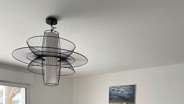 Een moderne woonkamer met een grijze bank, een televisie op een houten dressoir, een zwart ijzeren kroonluchter en een schilderij van een landschap.