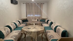 Ein gemütliches Wohnzimmer mit einer Sofagarnitur um einen zentralen Couchtisch, einem Fenster mit Vorhängen und einer wandmontierten Klimaanlage.