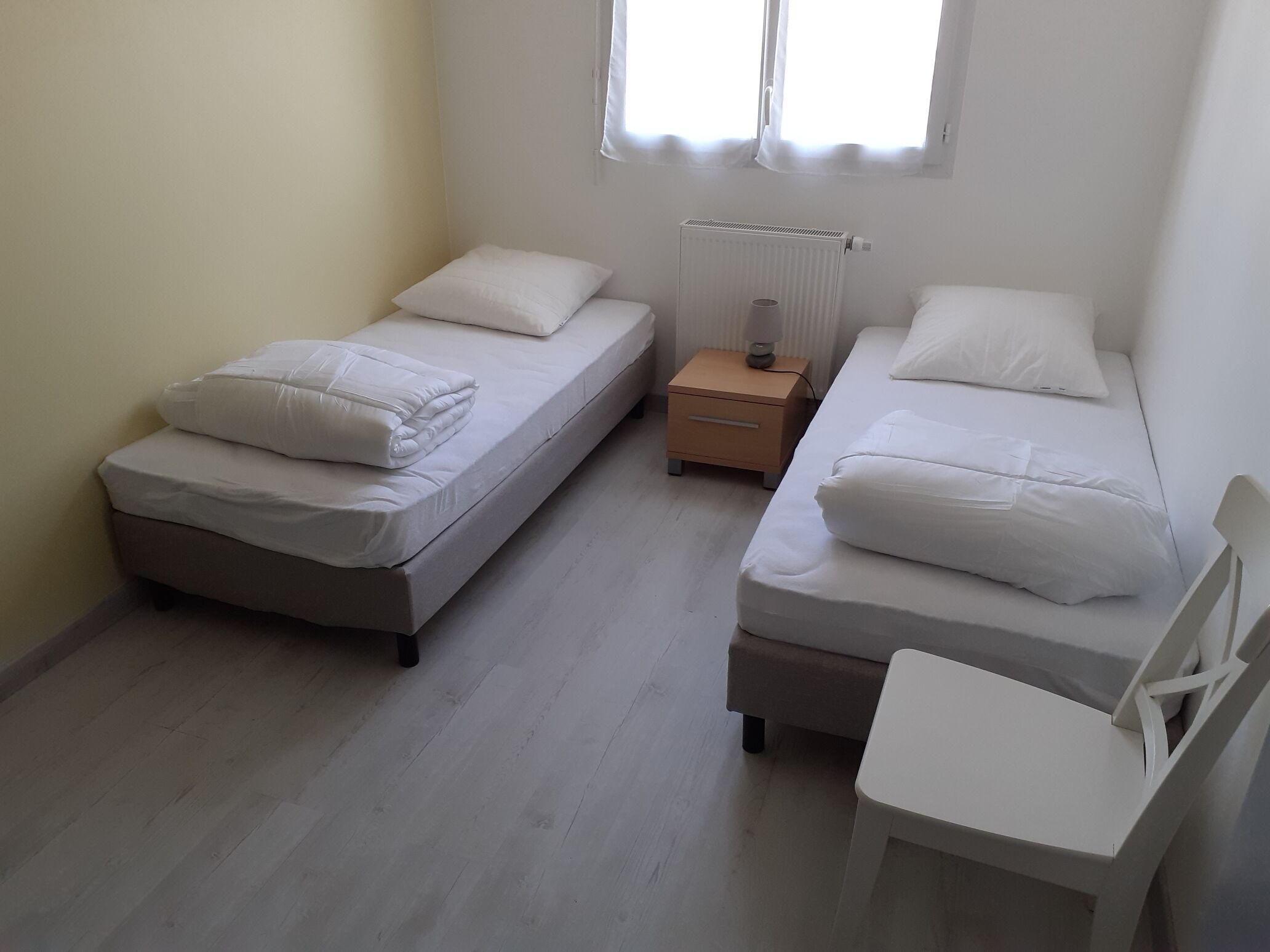 A small, clean room with two single beds, a nightstand, a chair, and a radiator.