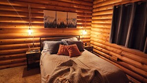 A cozy log cabin bedroom with a bed, two bedside tables, a chandelier, and a painting on the wall.