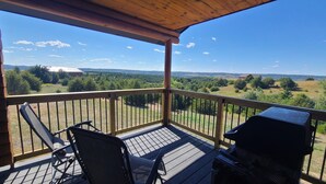 A wooden deck with two chairs and a grill, overlooking a rural landscape with trees and distant buildings.