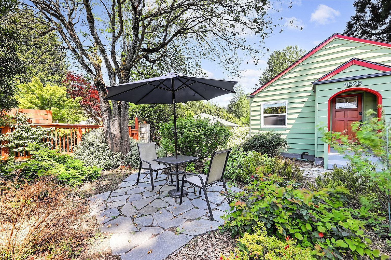 Een terras met een tafel, stoelen en een parasol, en een huis met een rode deur en het nummer 6526.