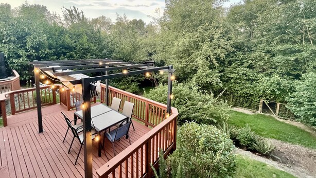 En træterrasse med et bord og stole, omgivet af frodig vegetation og træer.